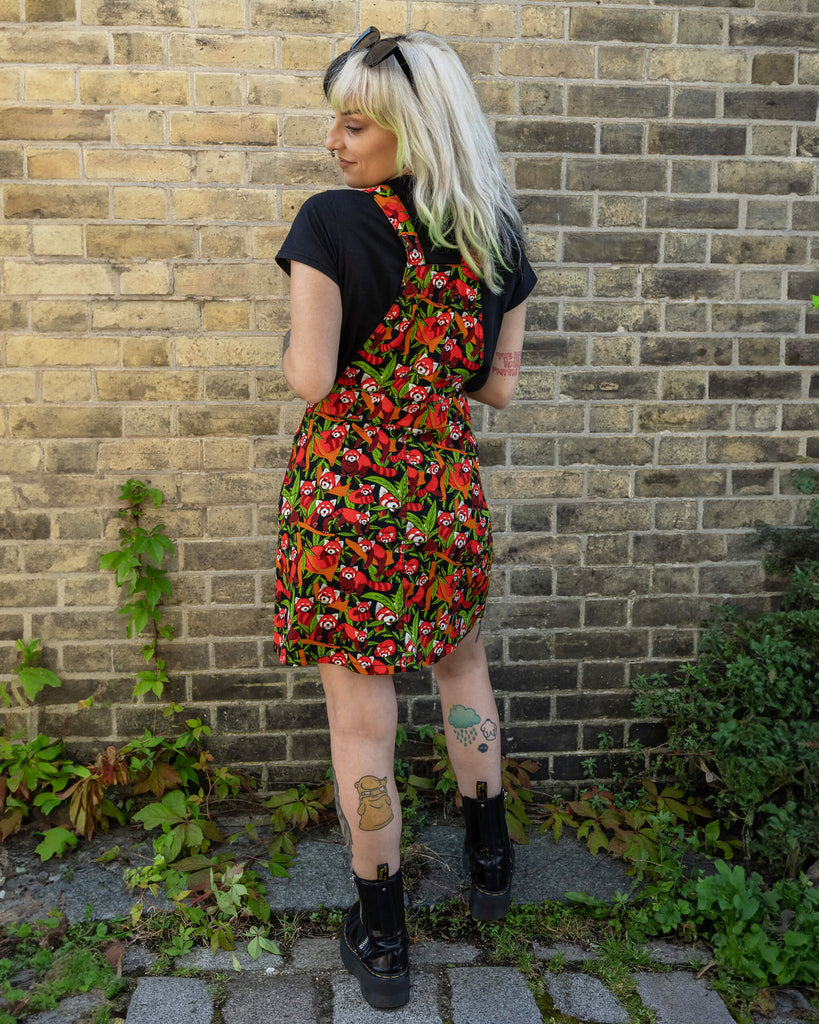 A woman with black and blonde hair is posing with her back to the camera, wearing a Run & Fly Red Panda pinafore. The pinafore displays a vibrant red panda and foliage design on a black background. She has her hands in the pinafore pockets and is standing against a brick wall.