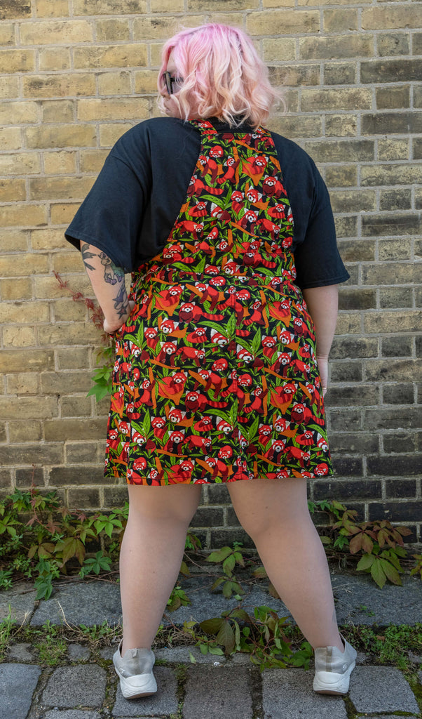 A model with pink hair and glasses is shown from the back, modelling a Run & Fly Red Panda pinafore. The pinafore features an eye-catching pattern of red pandas and green leaves on a black background. They are standing against a brick wall, highlighting the back details of the garment.