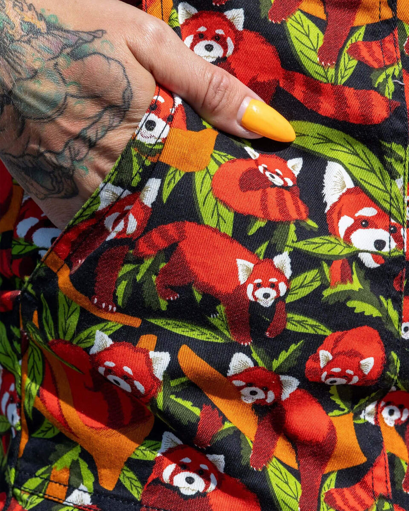 Close up of the Red Panda Stretch Twill Playsuit modelled by a femme tattooed model with long black hair with pink croc shoes and black sunglasses. She is outside along the beach with colourful beach huts behind her, she is facing forward showing her hand in the side pocket. The playsuit print is a black base covered with illustrated bamboo, branches and red pandas shown asleep, curled up, hanging off branches and eating the leaves.