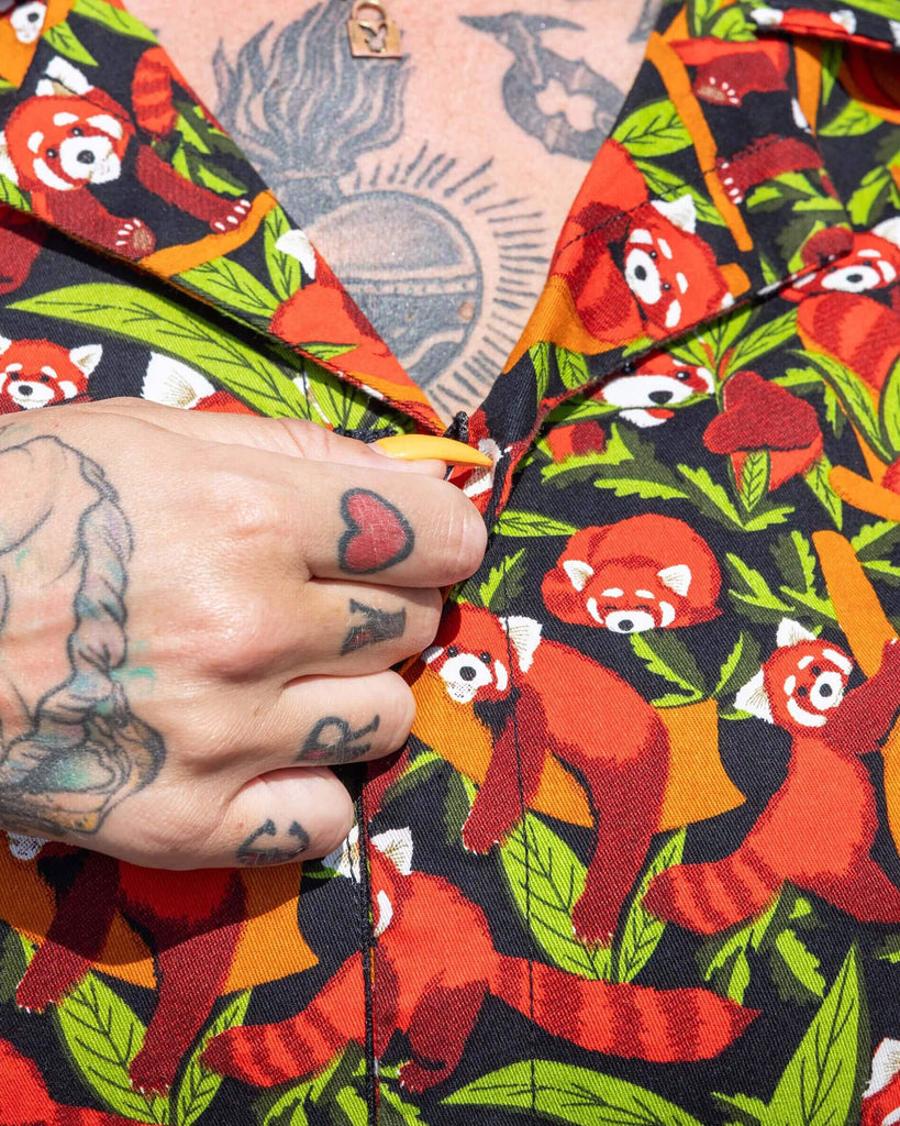 Close up of the Red Panda Stretch Twill Playsuit modelled by a femme tattooed model with long black hair with pink croc shoes and black sunglasses. She is outside along the beach with colourful beach huts behind her, she is facing forward showing the zip front feature. The playsuit print is a black base covered with illustrated bamboo, branches and red pandas shown asleep, curled up, hanging off branches and eating the leaves.