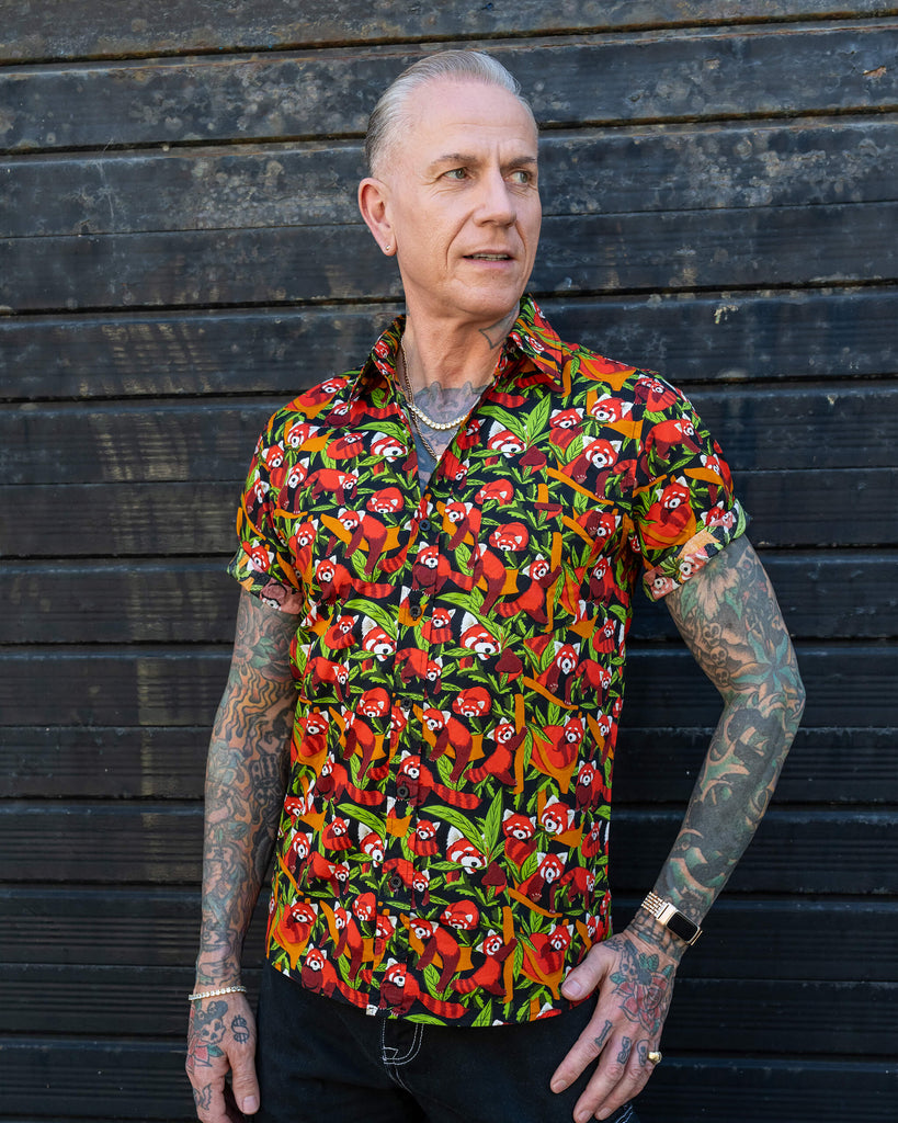 An older man with tattoos models a vibrant short-sleeve shirt featuring a red panda print. He stands against a dark wooden background, smiling and looking to the side.