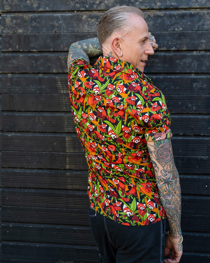 An older man with tattoos shows the side view of a bright short-sleeve shirt with a red panda pattern. He stands against a dark wooden background, turning to display the shirt's back while smiling.