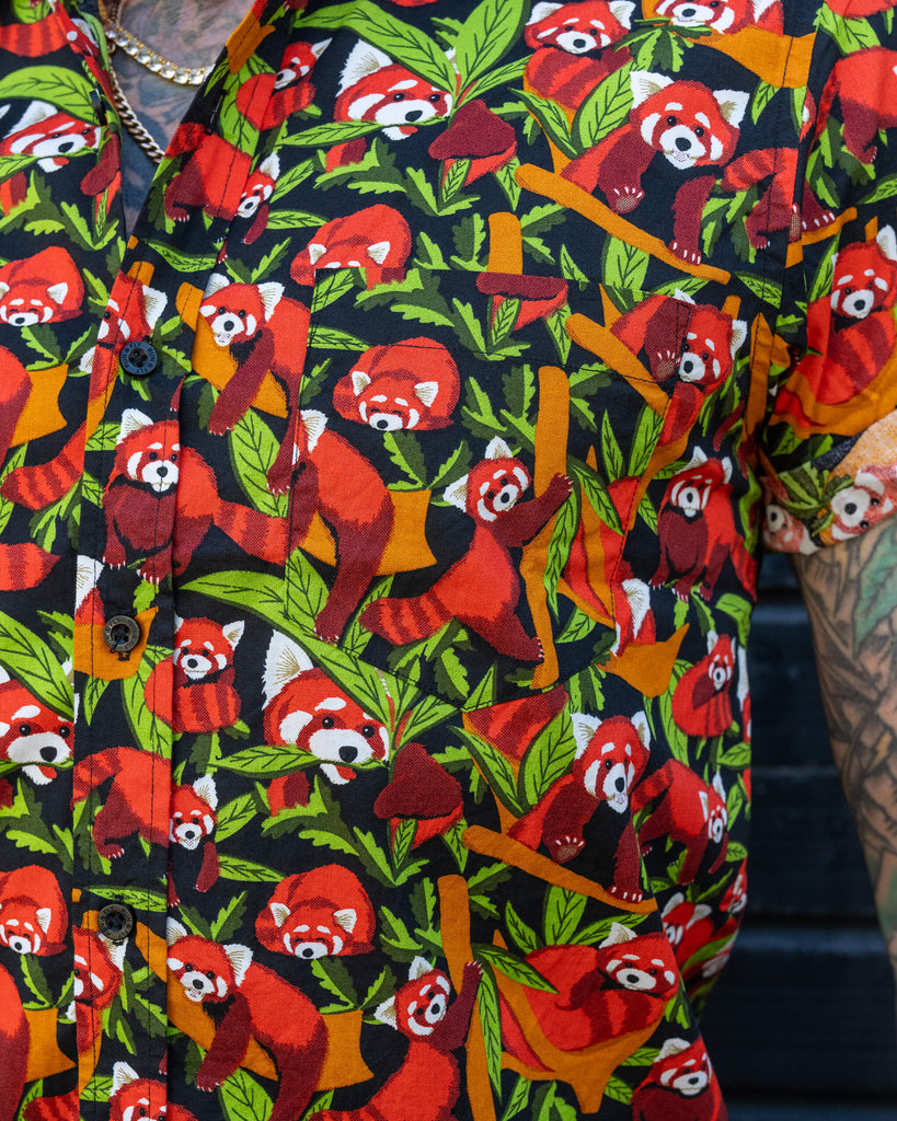 A close-up of a short-sleeve shirt with a red panda print, worn by an older man with tattoos. The design features multiple red pandas amidst green leaves on a dark background.
