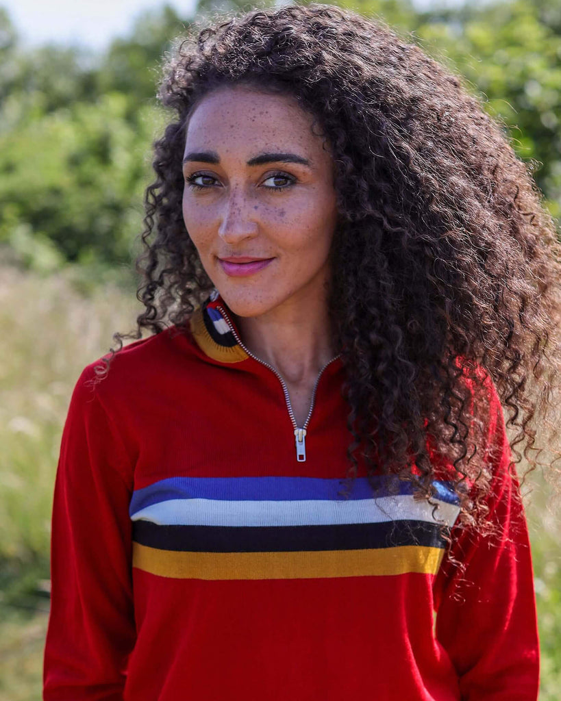 Model is looking at the camera in front of some greenery, whilst wearing dark shorts and the Red Retro Zip Neck Jumper.