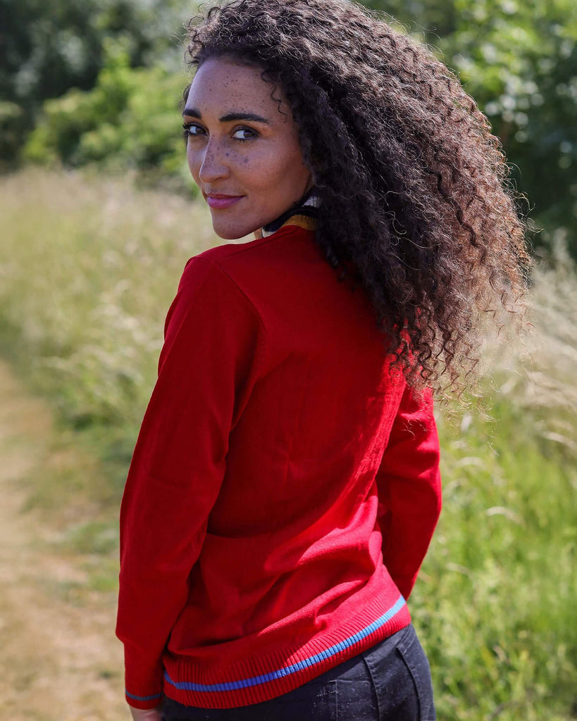 Model is looking over their shoulder towards the camera in front of some greenery, whilst wearing dark shorts and the Red Retro Zip Neck Jumper.
