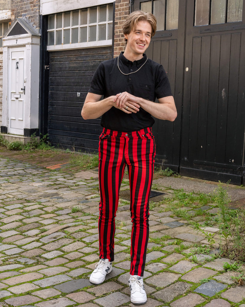 The model, dressed in Run & Fly's red and black striped skinny jeans, stands on a cobblestone street. Paired with a black polo and white trainers, the bold vertical stripes create a striking contrast. Run & Fly, a small ethical slow fashion brand, combines vintage-inspired style with modern flair.