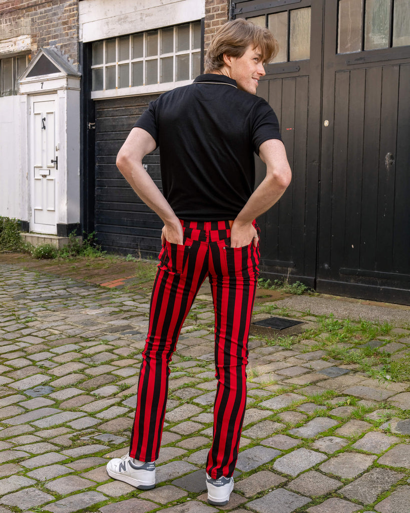 The model, now standing with hands in pockets, showcases the back of Run & Fly's red and black striped skinny jeans. The jeans hug the figure and feature two back pockets and a brown leather brand patch. The contrasting vertical stripes create a bold, striking look. Ethical slow fashion.