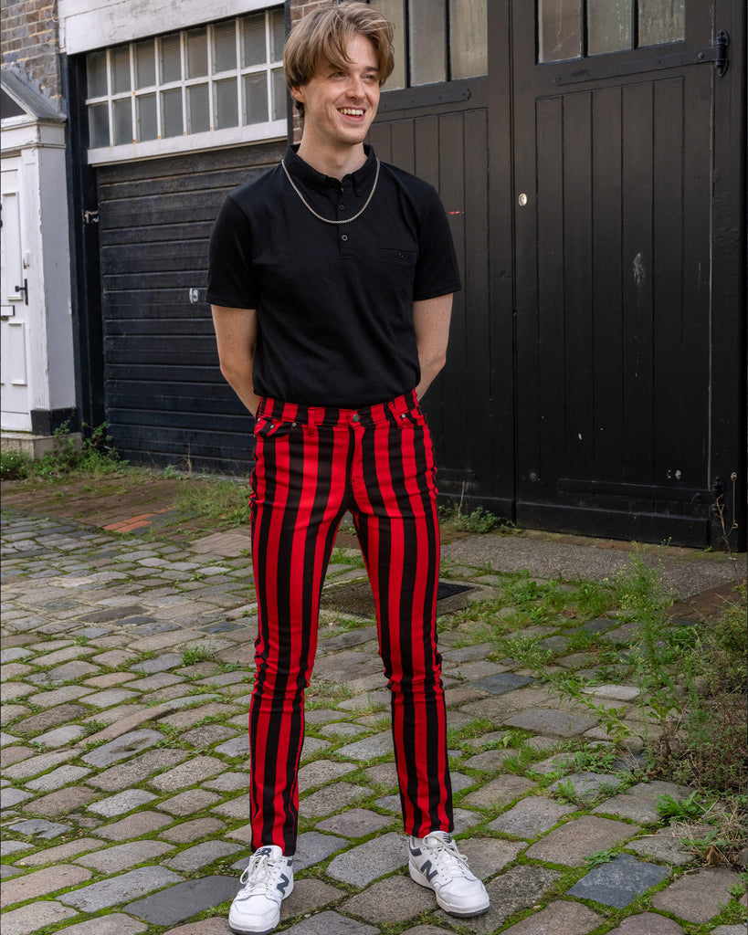 A man wearing Run & Fly red and black striped skinny jeans stands outdoors on cobblestone, styled with a black polo and white trainers. The jeans feature bold vertical stripes, and the brand's leather patch is visible on the waistband. He smiles, hands clasped together. Ethical slow fashion, unisex style.