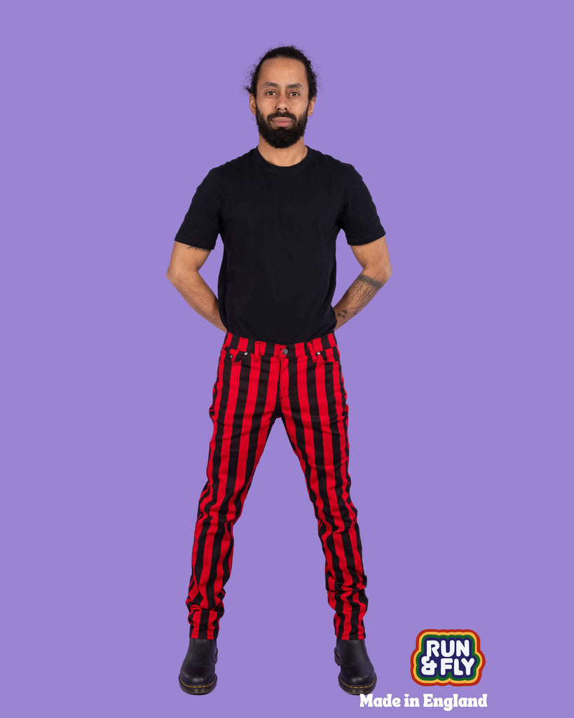 Richard, a hispanic male model with dark hair in a bun and a beard, is stood in a photography studio in Hove in front of a lilac backdrop wearing Black & Red 1" Striped Mid Rise Stretch Skinny Jeans with a black t shirt and black shoes. Richard is smiling and facing the camera with his arms behind his back and legs slightly apart. Run & Fly logo is at the corner or the photo with 'Made in England' written underneath.