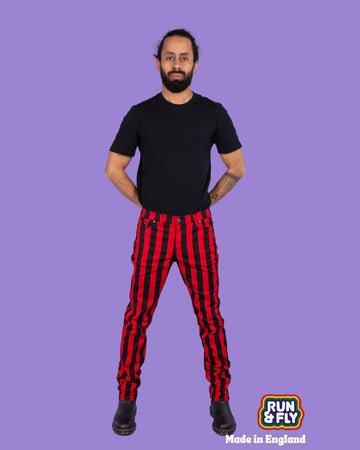 Richard, a hispanic male model with dark hair in a bun and a beard, is stood in a photography studio in Hove in front of a lilac backdrop wearing Black & Red 1" Striped Mid Rise Stretch Skinny Jeans with a black t shirt and black shoes. Richard is smiling and facing the camera with his arms behind his back and legs slightly apart. Run & Fly logo is at the corner or the photo with 'Made in England' written underneath.