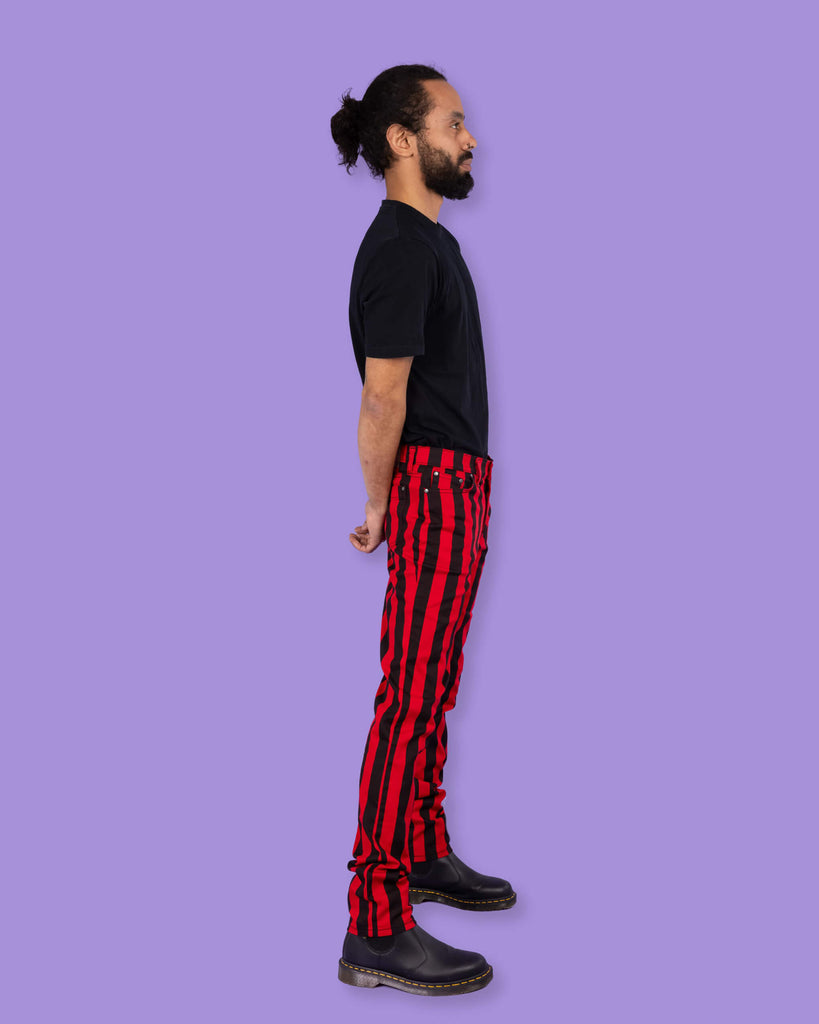 Richard, a hispanic male model with dark hair in a bun and a beard, is stood in a photography studio in Hove in front of a lilac backdrop wearing Black & Red 1" Striped Mid Rise Stretch Skinny Jeans with a black t shirt and black shoes. Richard is stood sideways with his arms behind his back.