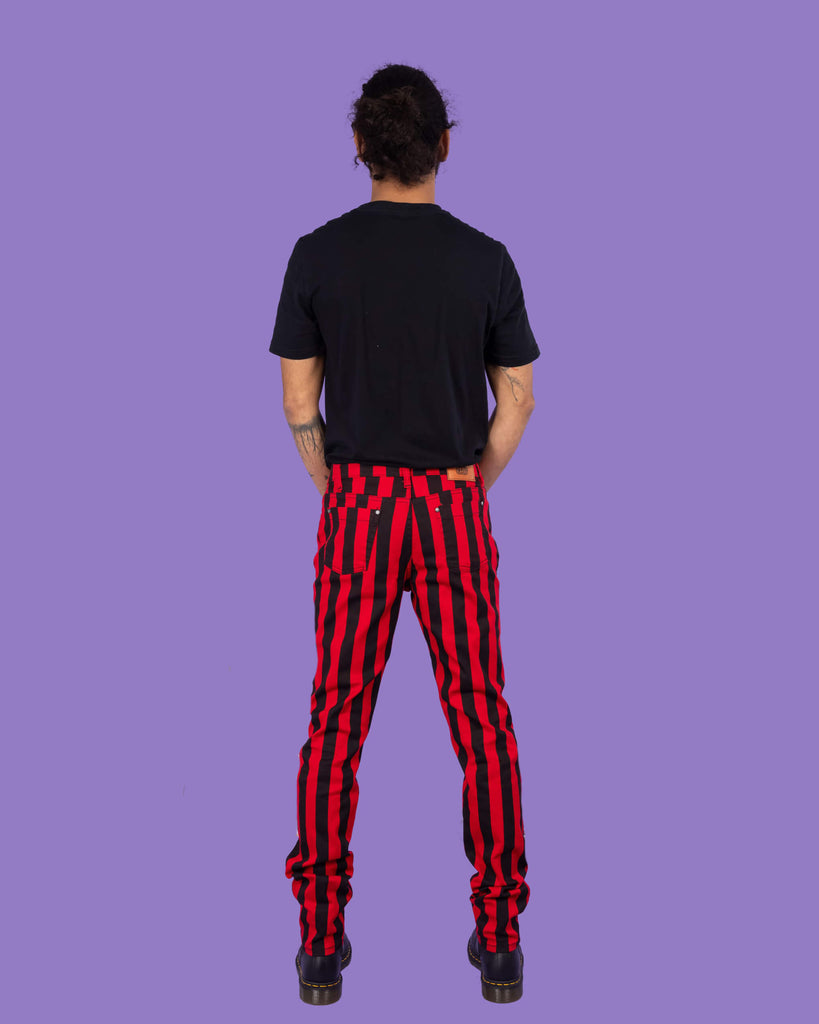 Richard, a hispanic male model with dark hair in a bun and a beard, is stood in a photography studio in Hove in front of a lilac backdrop wearing Black & Red 1" Striped Mid Rise Stretch Skinny Jeans with a black t shirt and black shoes. Richard is posing with his back to the camera and arms in front of him.