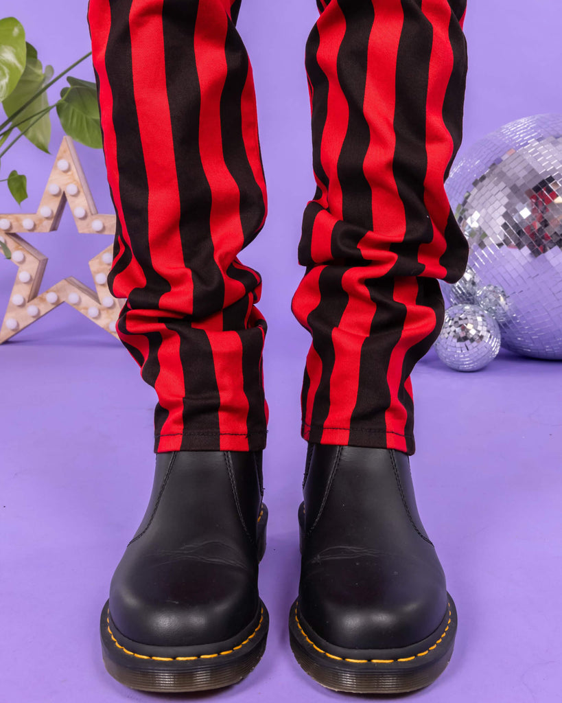 Close up of Richard wearing the Black & Red 1" Striped Mid Rise Stretch Skinny Jeans with black shoes. The background of the photo is lilac and there are disco balls and a star shaped light behind him. The photo is cropped at the knees.