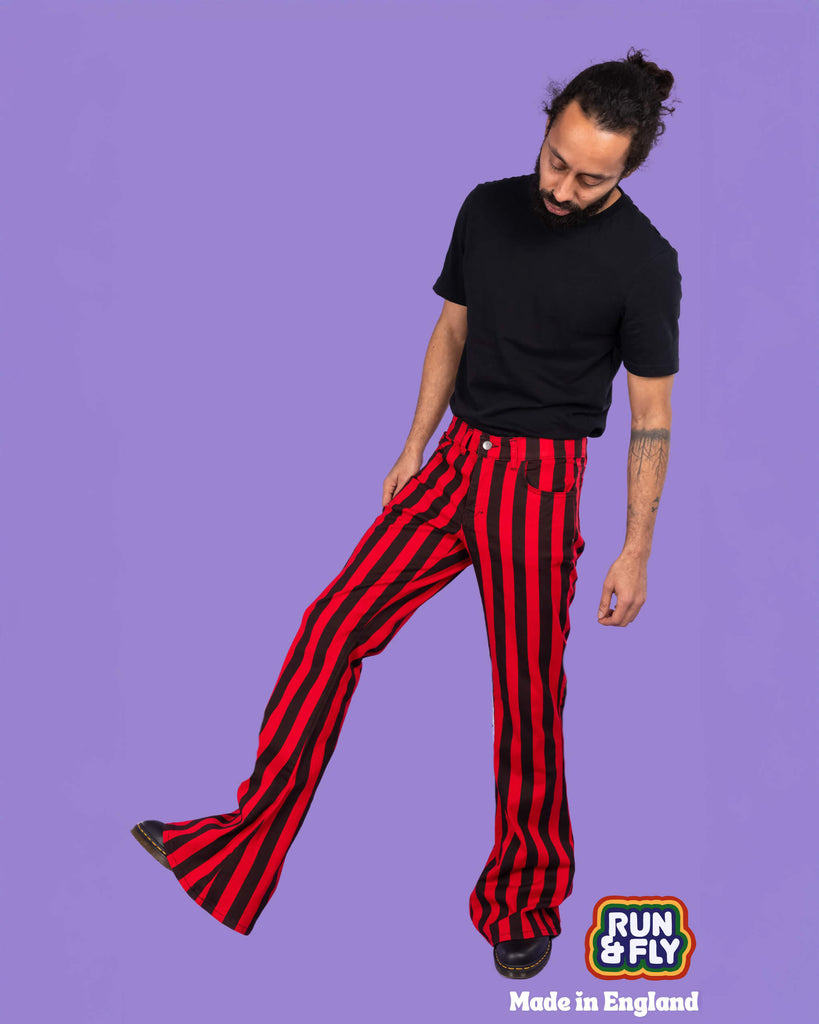 Richard, a hispanic male model with dark hair in a bun and a beard, is stood in a photography studio in Hove in front of a lilac backdrop wearing Red & Black Striped Bell Bottom Stretch Super Flares with a black t shirt and black shoes. Richard is facing the camera with one leg turned out to the side and lifted in the air. He is looking down with his arms down by his sides. Run & Fly logo is at the corner or the photo with 'Made in England' written underneath.