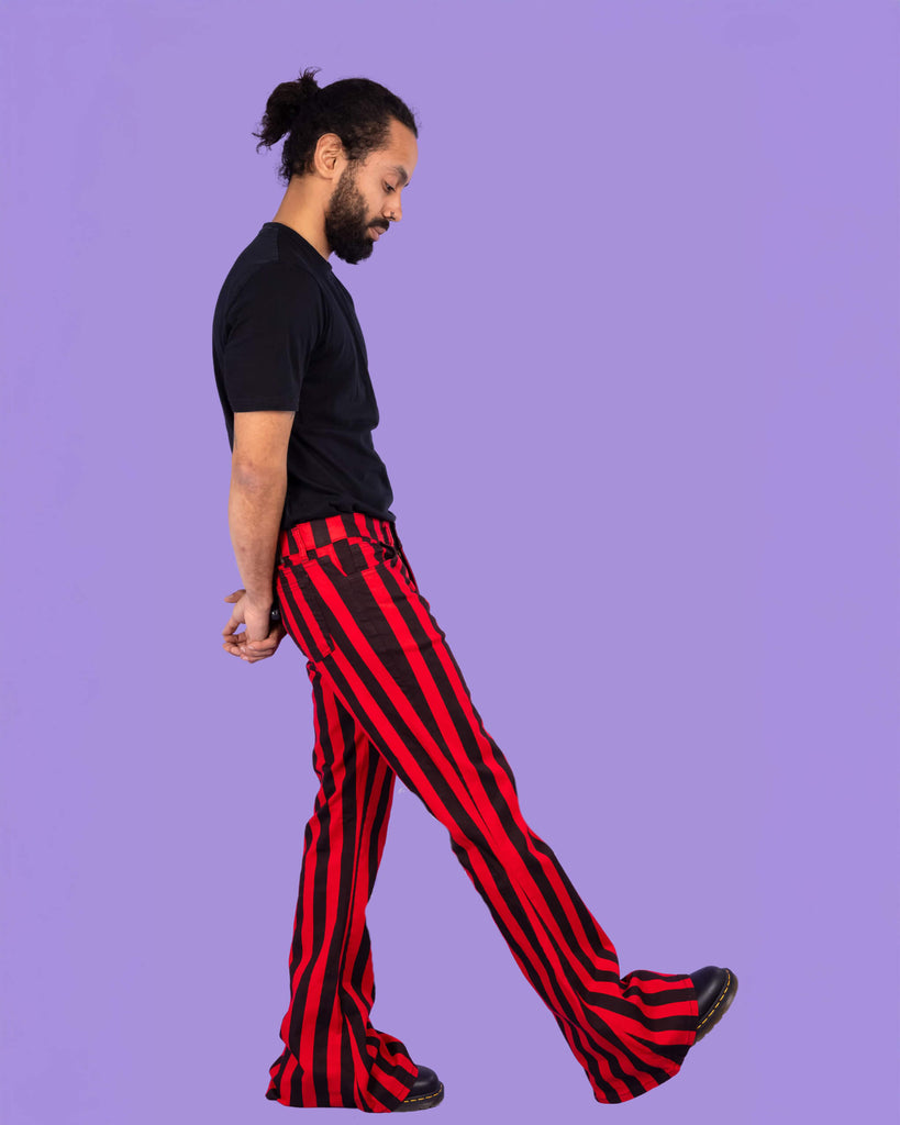 Richard, a hispanic male model with dark hair in a bun and a beard, is stood in a photography studio in Hove in front of a lilac backdrop wearing Red & Black Striped Bell Bottom Stretch Super Flares with a black t shirt and black shoes. Richard is stood sideways with one leg outstretched in front of him and is looking down with his arms behind him.