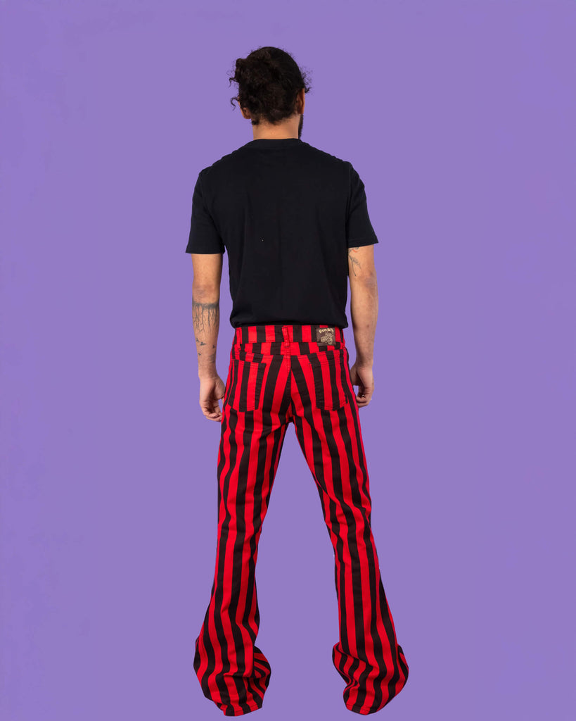 Richard, a hispanic male model with dark hair in a bun and a beard, is stood in a photography studio in Hove in front of a lilac backdrop wearing Red & Black Striped Bell Bottom Stretch Super Flares with a black t shirt and black shoes. Richard is posing with his back to the camera and his arms down by his side.