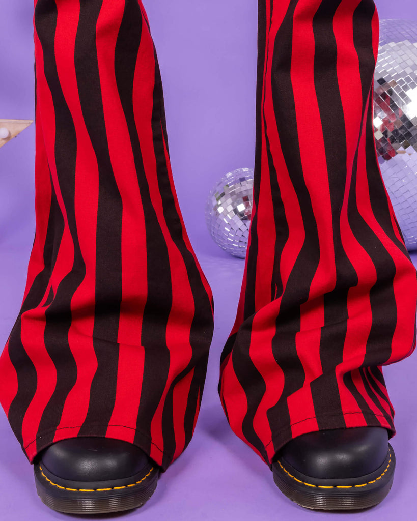 Close up of Richard wearing the Red & Black Striped Bell Bottom Stretch Super Flares with black shoes. The background of the photo is lilac and there are disco balls behind him. The photo is cropped at the knees.