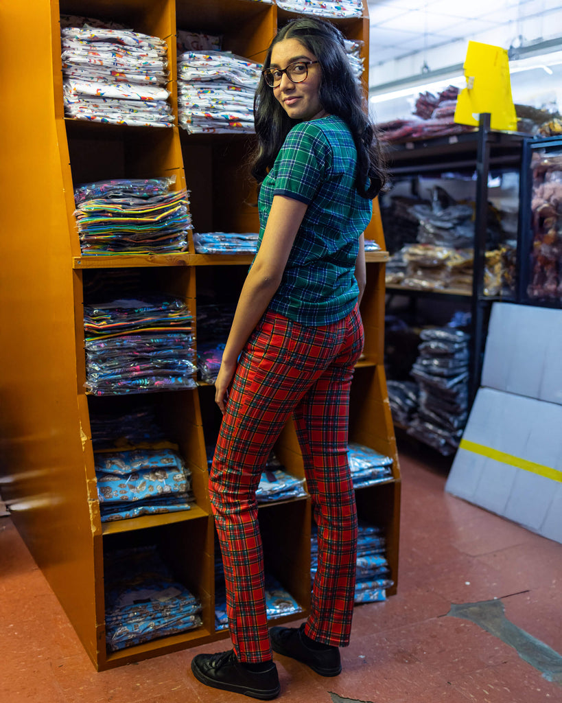 Model is stood in our warehouse wearing the mid rise red tartan stretch skinny jeans with a green tartan tshirt and black trainers. They are facing away from the camera to highlight the back details. Photo is cropped from the waist down.