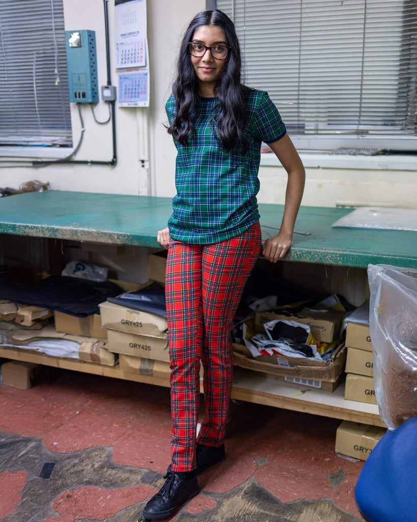 Model is stood in our warehouse area wearing the mid rise red tartan stretch skinny jeans with a green tartan tshirt and black trainers. They are facing towards the camera leaning against a desk with both hands behind their back and smiling.