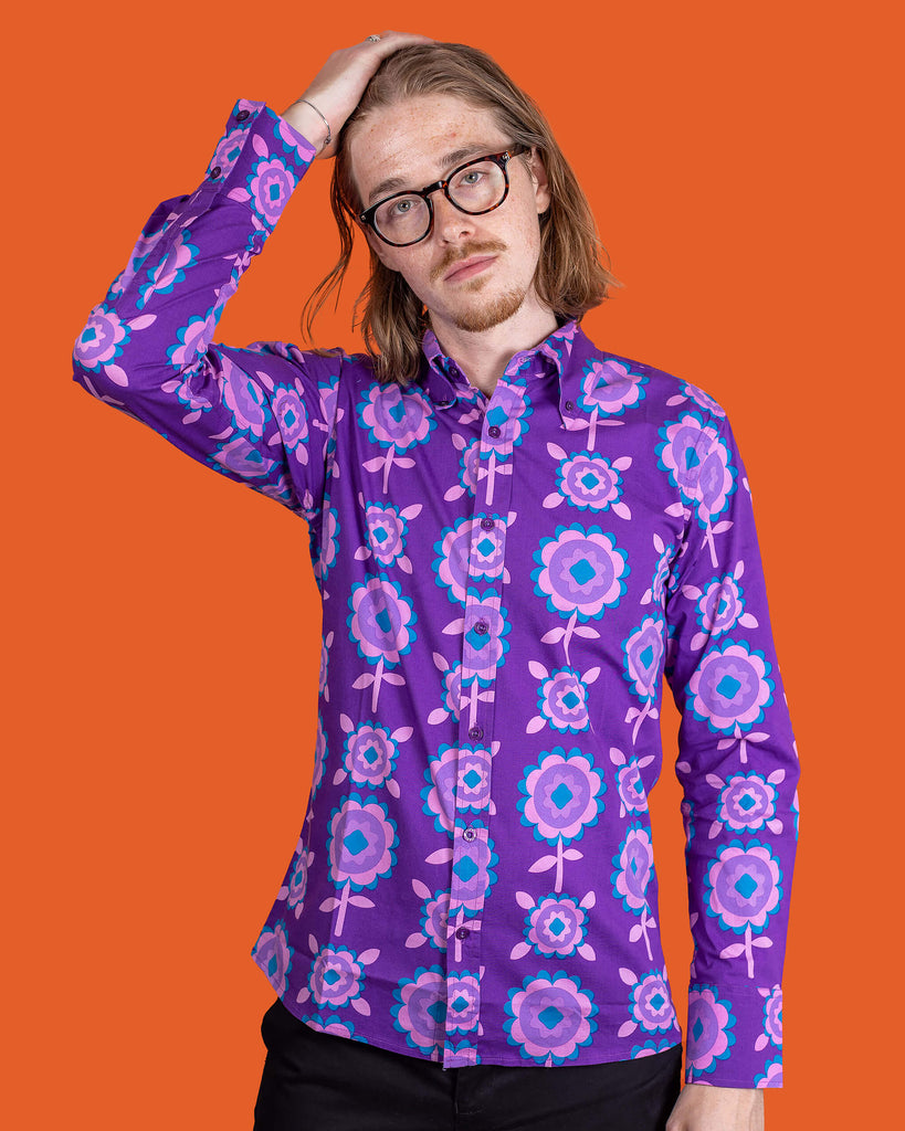 Jack, a male model with shoulder length hair and glasses is stood in front of an orange backdrop at a photography studio in Hove wearing Purple Retro Flowers Long Sleeve Shirt with black trousers. Jack is posing toward the camera with one hand in his hand and other down by his side. The photo is cropped at the knees.