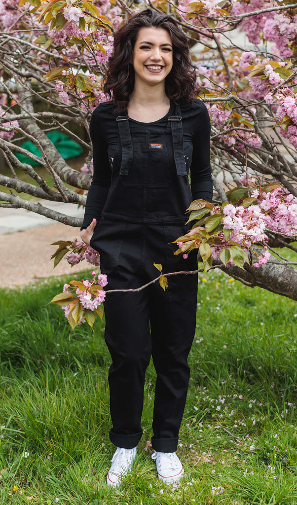 Sophie smiling and standing in front of a tree wearing the Black Stretch Denim Dungarees with a long sleeve black t-shirt and white trainers