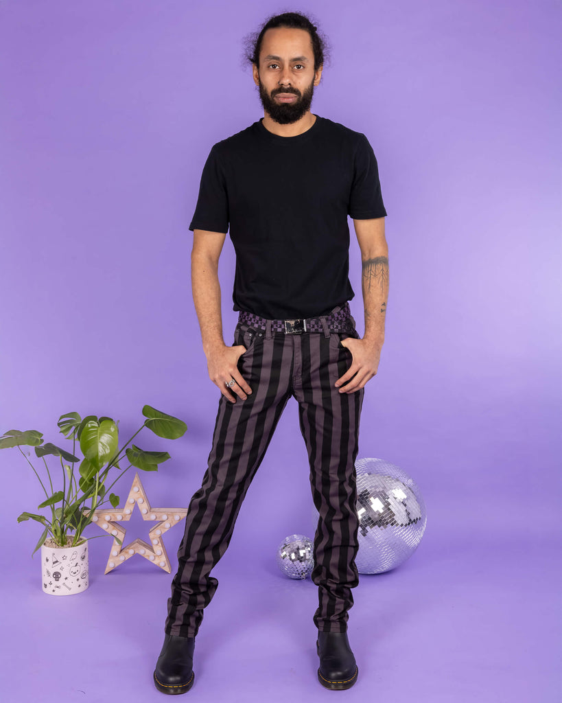 Richard, a hispanic male model with dark hair in a bun and a beard, is stood in a photography studio in Hove in front of a lilac backdrop wearing Black & Grey 1" Striped Mid Rise Stretch Skinny Jeans with a purple and black checkered belt, a black t shirt and black shoes. Richard is posing facing the camera with one leg slightly out to the side and his thumbs are resting in the front pockets of the jeans. Run & Fly logo is at the corner or the photo with 'Made in England' written underneath.