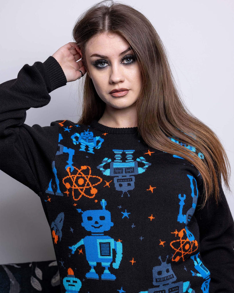 Charlotte is stood in front of a grey sofa wearing the robot jumper with black stone wash flares. She is facing forward with one hand in her hair and other resting by her side and is posing at the camera. Photo is cropped from the thighs up. The robot jumper is a black base featuring multiple blue robots with orange, blue stars and chemical symbols with plain black arms and back.