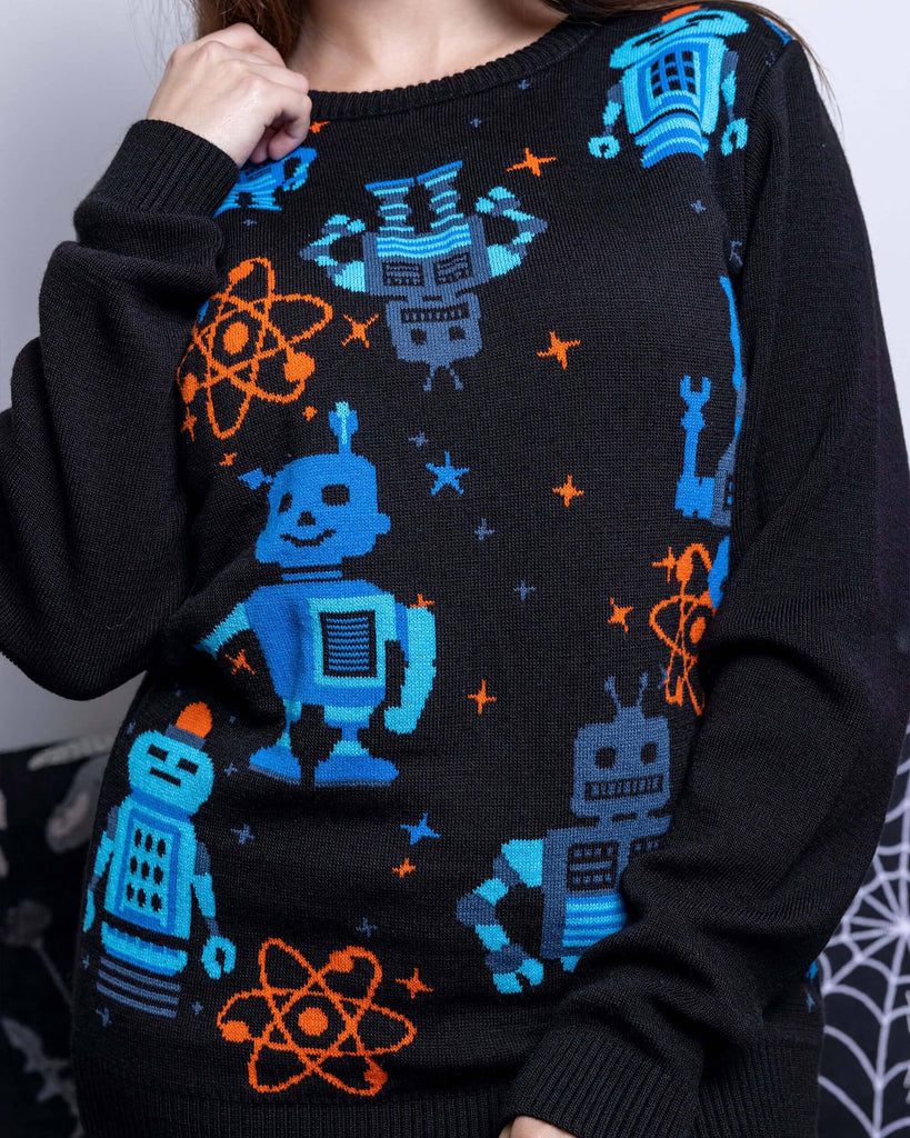 Charlotte is stood in front of a grey sofa wearing the robot jumper with black stone wash flares. She is facing forward with one hand resting on her collarbone and the other hand resting by her side. Photo is cropped from the neck to hips. The robot jumper is a black base featuring multiple blue robots with orange, blue stars and chemical symbols with plain black arms and back.