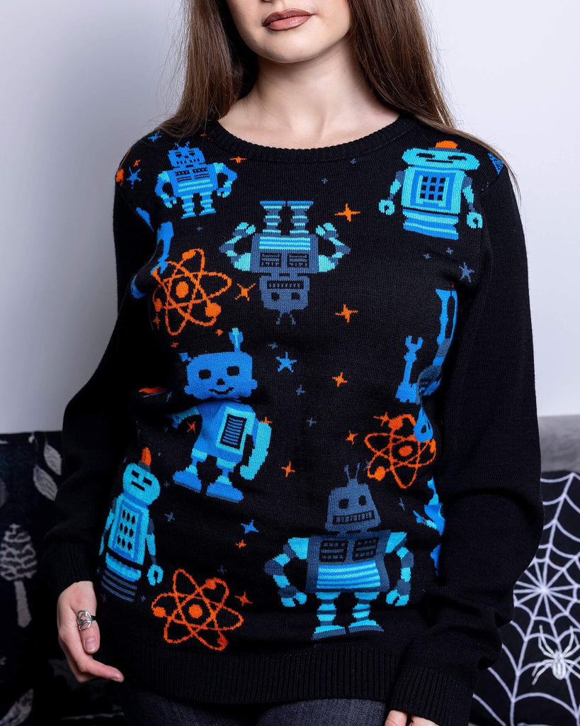 Charlotte is stood in front of a grey sofa wearing the robot jumper with black stone wash flares. She is facing forward with both hands resting by her side and smiling to camera. Photo is cropped from the thighs up. The robot jumper is a black base featuring multiple blue robots with orange, blue stars and chemical symbols with plain black arms and back.