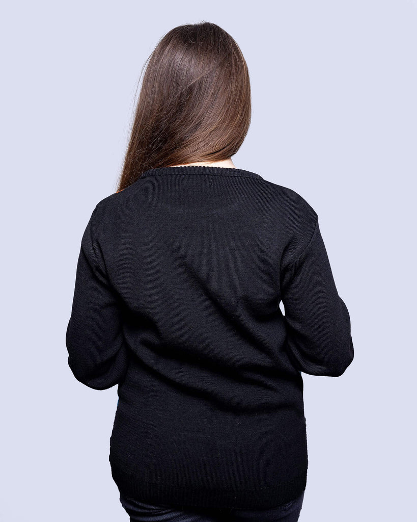 Charlotte is stood in front of a grey sofa wearing the robot jumper with black stone wash flares. She is facing away from the camera with both arms resting by her sides. Photo is cropped from the thighs up. The robot jumper is a black base featuring multiple blue robots with orange, blue stars and chemical symbols with plain black arms and back.