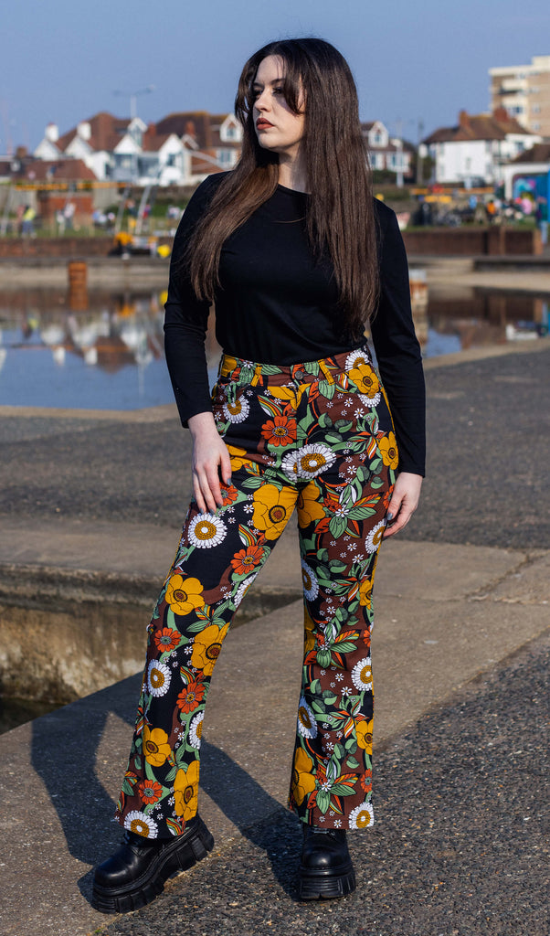 Alice with mid length brown hair is wearing the 70s black floral high waisted bell bottom flares with a long sleeve black tshirt and black boots. She is facing forward posing with one leg bent outwards to highlight the trouser shape whilst looking off to the left.