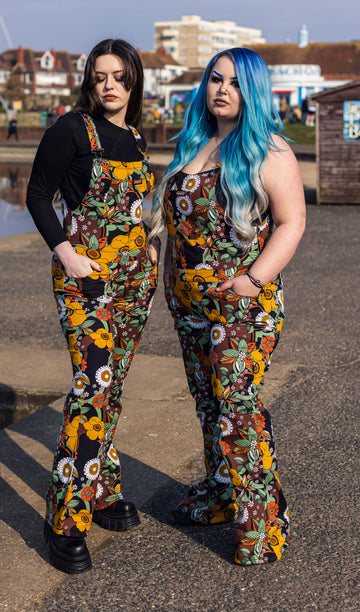 Alice and Azariah stood near a beach park area wearing the 70's black floral stretch skinny flared dungarees. They are both facing forward angled inward to highlight the flared shape. Alice has mid length brown hair and is wearing a long sleeve black tshirt underneath with black platform boots. Azariah has long blue gradient hair and is wearing a strappy black crop top underneath with black boots.