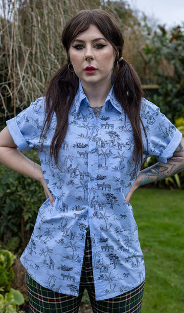 Flo is a femme dark haired model with tattoos she  is stood in front of a garden wearing the blue safari short sleeve shirt with tartan flares.