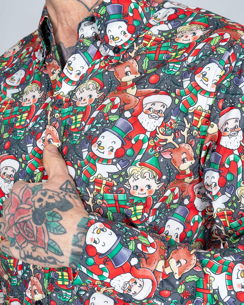 A close-up of a colourful long-sleeved Christmas shirt by Run & Fly, featuring retro-style designs of Santa Claus, elves, snowmen, reindeer, and festive gifts on a dark background. The shirt is buttoned up, showcasing the fine details of the quirky, vintage-inspired print. Ethical slow fashion meets holiday spirit in this standout piece.