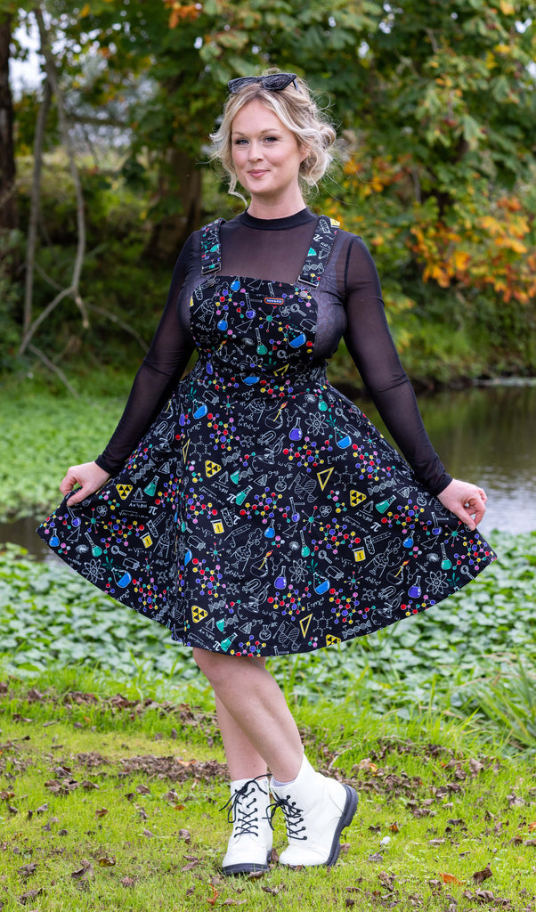 Karina is stood outside by a river wearing School of Science Flared Pinafore Dress with a long sleeve black mesh top underneath, and white boots. The back dress has an all over print of various chemistry equipment in white, yellow, green, red, yellow and purple. Karina is posing to the camera with one leg bent and is holding the skirt of the pinafore out to her sides.