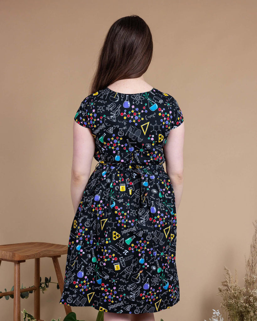 Charlotte is stood in front of a beige backdrop with various plants and disco balls behind her. She is wearing School of Science Belted Tea Dress with Pockets with black boots. The dress has a black background colour with an all over print of various chemistry equipment in white, yellow, green, red, yellow and purple. Charlotte is stood with her back to the camera with her arms by her sides.