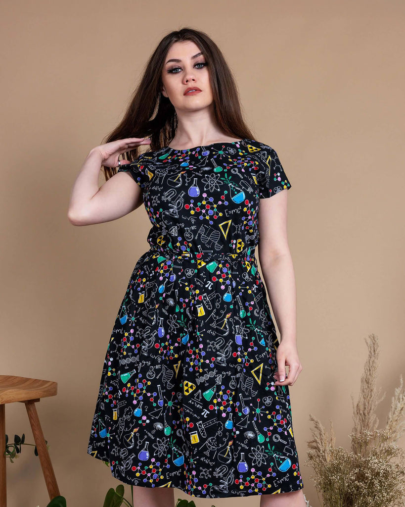 Charlotte is stood in front of a beige backdrop with various plants and disco balls behind her. She is wearing School of Science Belted Tea Dress with Pockets with black boots. The dress has a black background colour with an all over print of various chemistry equipment in white, yellow, green, red, yellow and purple. Charlotte is posing to the camera flipping her hair.
