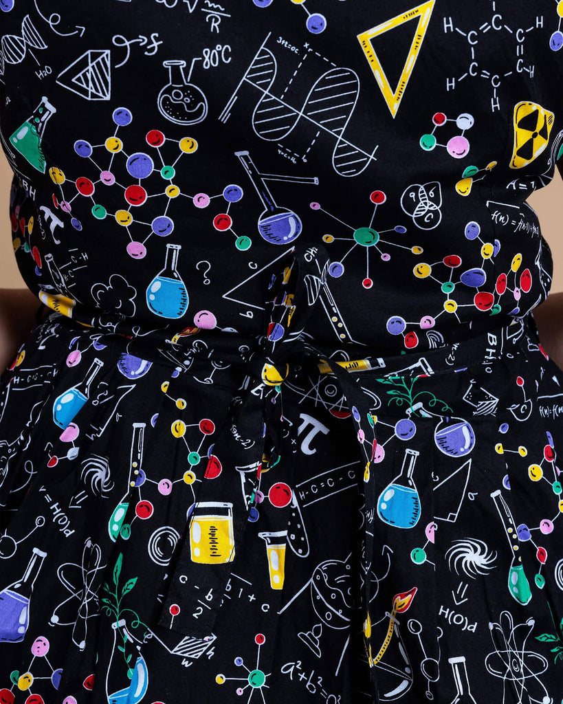 close up of the School of Science Belted Tea Dress with Pockets. The dress has a black background colour with an all over print of various chemistry equipment in white, yellow, green, red, yellow and purple.