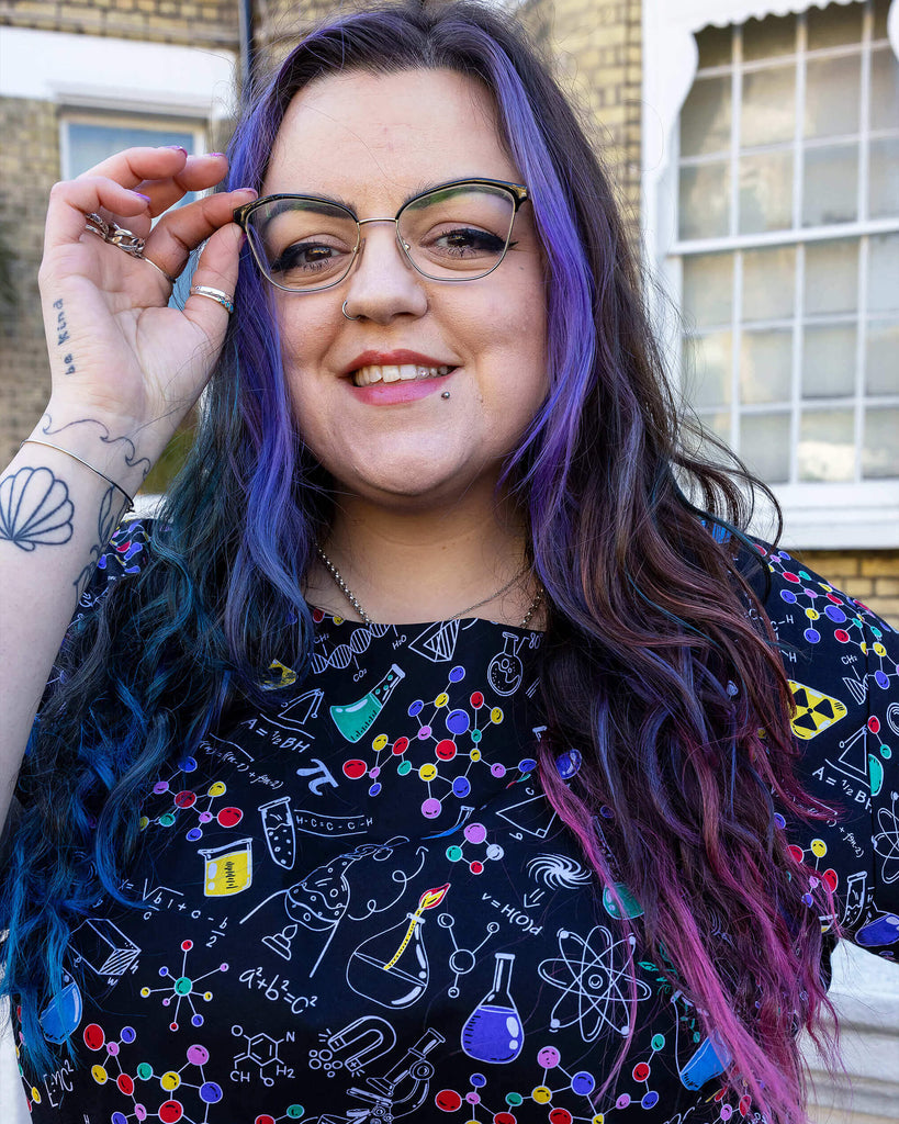 close up of Luisa wearing School of Science Belted Tea Dress with Pockets. The dress has a black background colour with an all over print of various chemistry equipment in white, yellow, green, red, yellow and purple. Luisa is stood outside in Hove smiling at the camera with one hand on her hip and other holding her glasses.