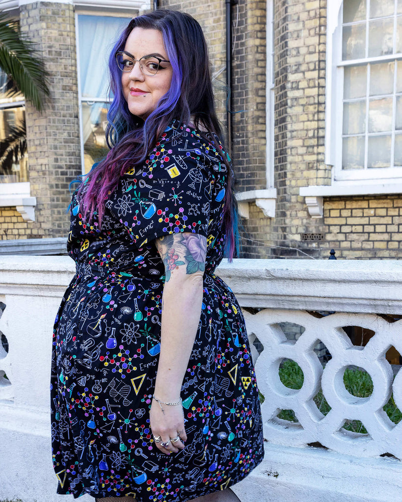 Luisa, a femme model with purple and blue hair and glasses, is stood outside a town house in Hove wearing School of Science Belted Tea Dress with Pockets with fishnet tights and black boots. The dress has a black background colour with an all over print of various chemistry equipment in white, yellow, green, red, yellow and purple. Luisa is facing away from the camera with one leg in front of the other and is looking back at the camera.