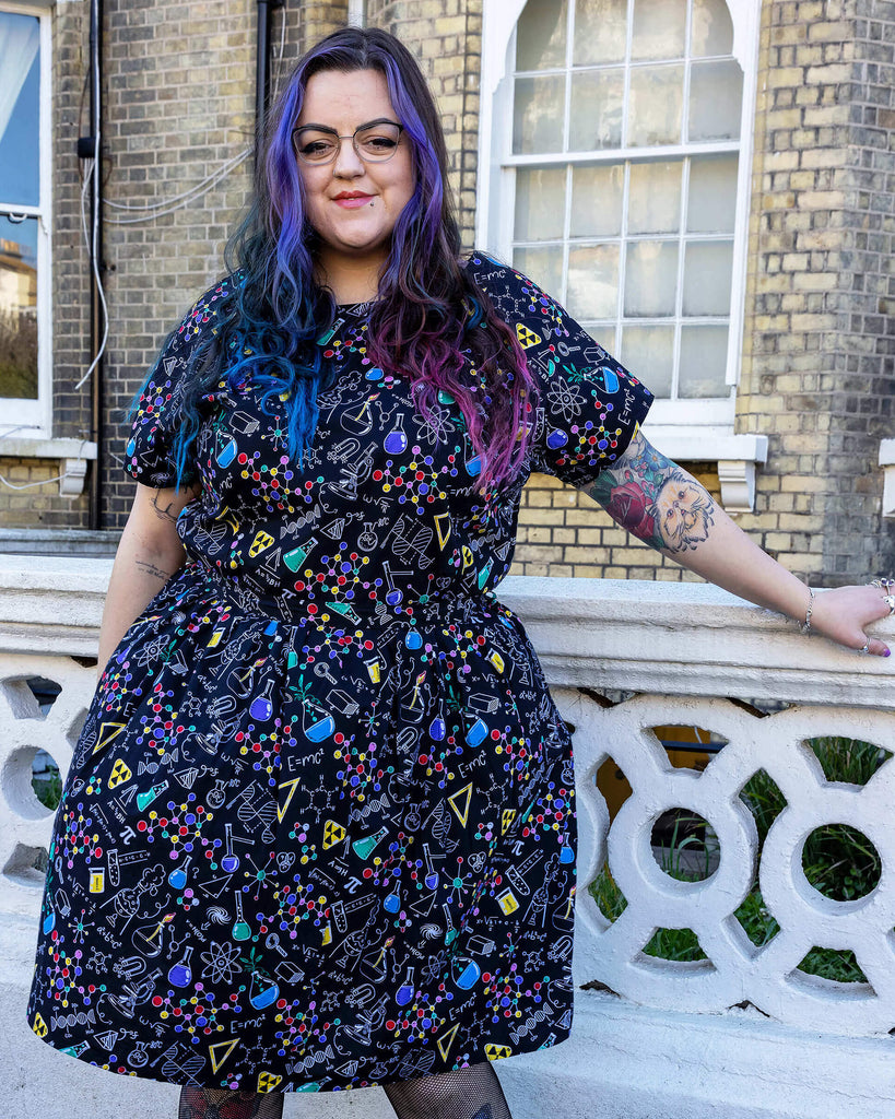 Luisa, a femme model with purple and blue hair and glasses, is stood outside a town house in Hove wearing School of Science Belted Tea Dress with Pockets with fishnet tights and black boots. The dress has a black background colour with an all over print of various chemistry equipment in white, yellow, green, red, yellow and purple. Luisa is facing the camera with one hand resting on a concrete railing and other arm resting down by her side.