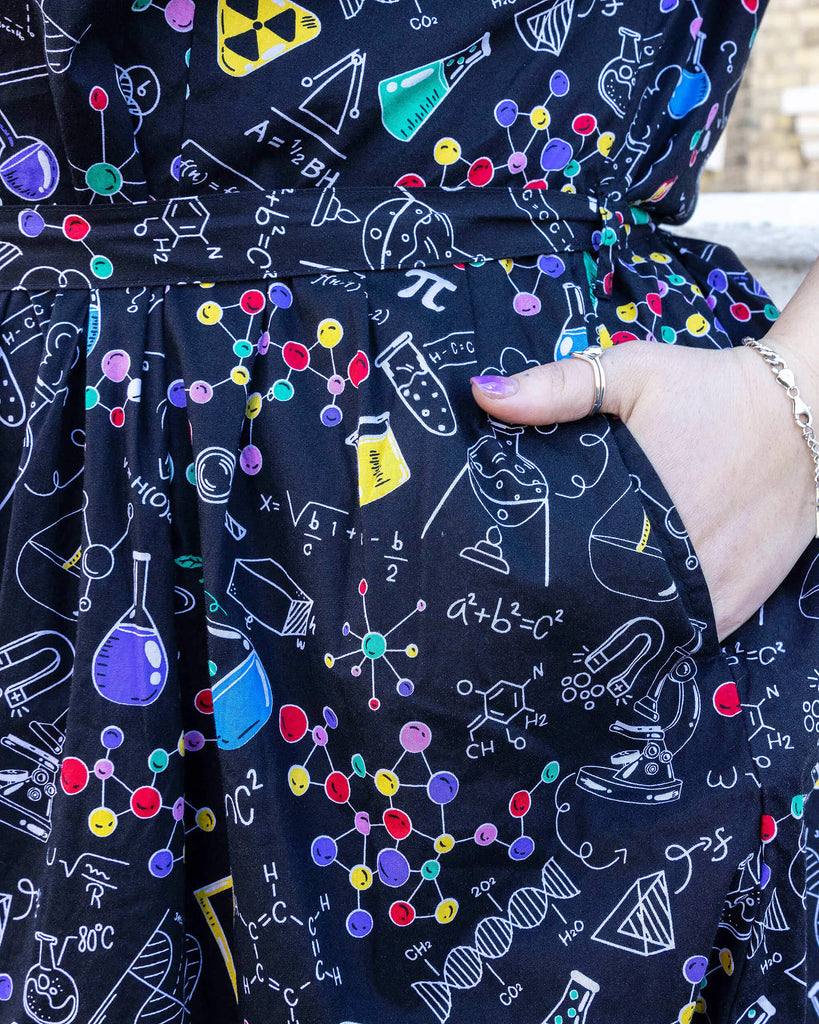 close up of model's hand in the pocket of School of Science Belted Tea Dress with Pockets. The dress has a black background colour with an all over print of various chemistry equipment in white, yellow, green, red, yellow and purple. The matching fabric belt is tied around the model's waist.