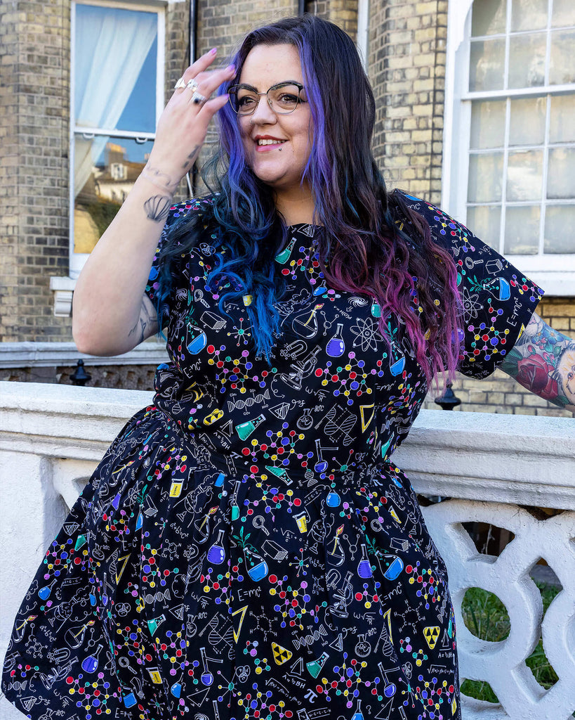 Luisa, a femme model with purple and blue hair and glasses, is stood outside a town house in Hove wearing School of Science Belted Tea Dress with Pockets with fishnet tights and black boots. The dress has a black background colour with an all over print of various chemistry equipment in white, yellow, green, red, yellow and purple. Luisa is posing looking off to one side with her hand in her hair.