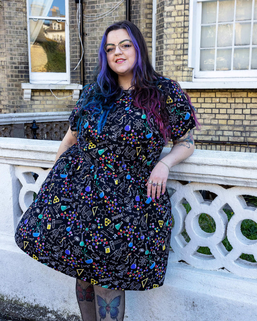Luisa, a femme model with purple and blue hair and glasses, is stood outside a town house in Hove wearing School of Science Belted Tea Dress with Pockets with fishnet tights and black boots. The dress has a black background colour with an all over print of various chemistry equipment in white, yellow, green, red, yellow and purple. Luisa is leant against a wall posing at the camera.