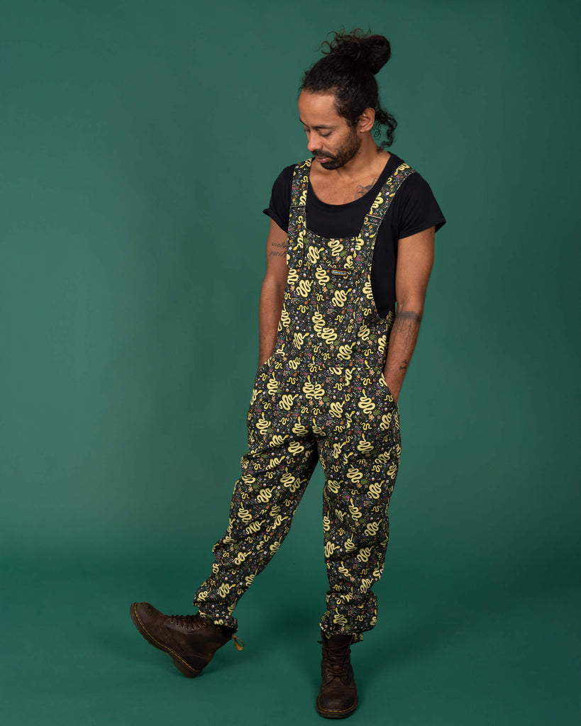 Full-body image of a model in Run & Fly serpent garden dungarees. The yellow snake and floral print is spread across the entire garment, paired with brown boots for a casual look. Small, ethical UK-based brand known for sustainable, gender-neutral fashion with playful designs.