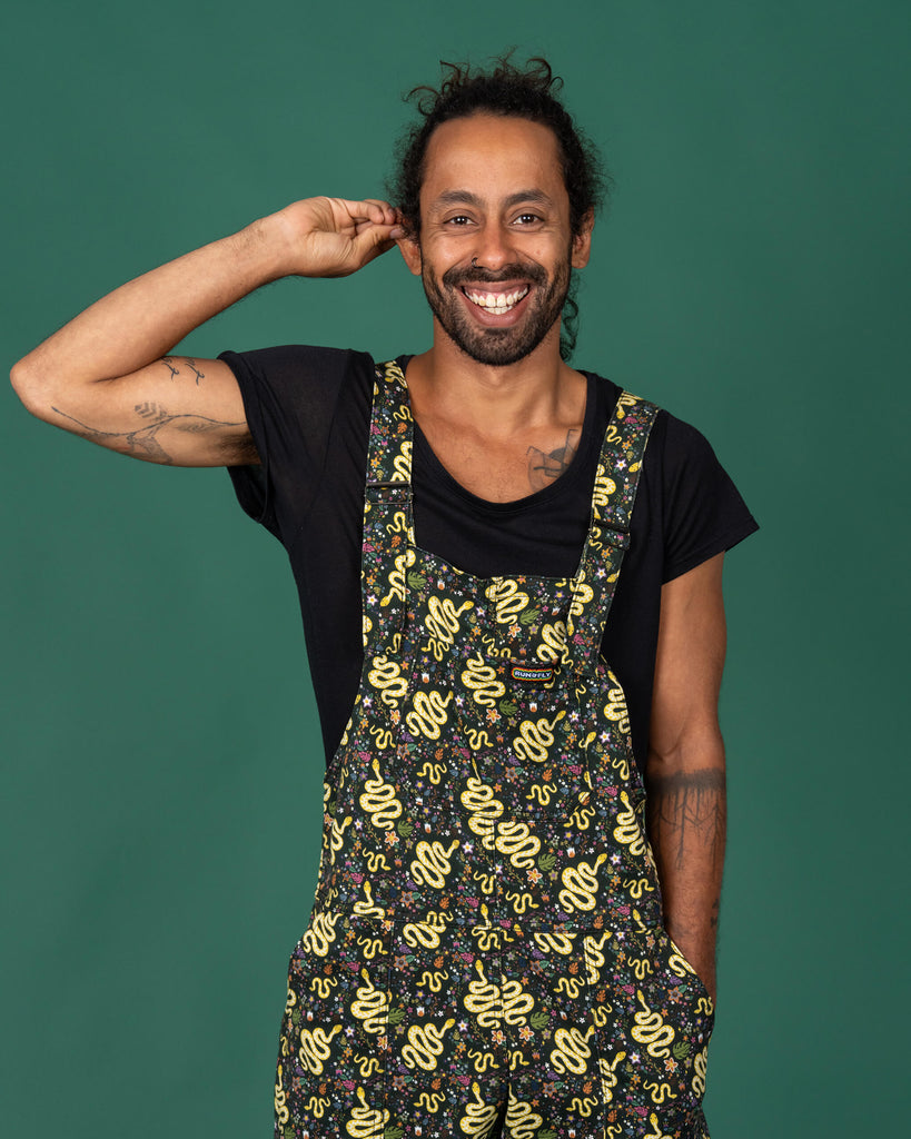 Smiling model wearing Run & Fly serpent garden dungarees, front-facing with hands in pockets. The dungarees feature a yellow snake and floral print against a black background, standing out as a statement piece. Ethical slow fashion brand known for fun, bold designs and comfortable fits