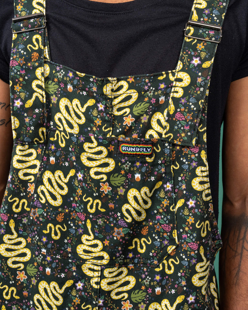 Close-up of the chest pocket of Run & Fly's serpent garden dungarees, with the brand's logo prominently featured. The yellow serpents weave through a floral and leafy print, creating a bold and intricate design. This ethical, slow fashion piece highlights the brand’s commitment to eco-friendly practices.