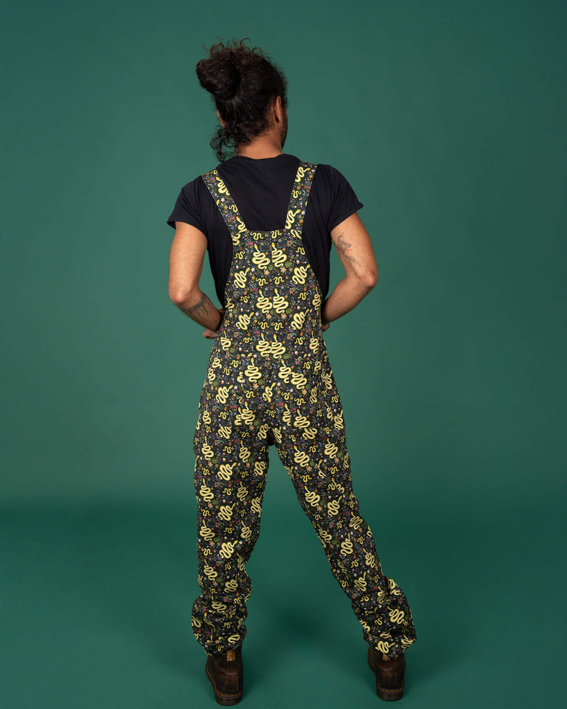 Full back view of a model wearing Run & Fly's serpent garden dungarees. The yellow snake and botanical print stands out on the black fabric, complemented by the model's casual pose. The dungarees' adjustable straps and relaxed fit make them ideal for comfort and sustainability. Small ethical brand, UK-made.