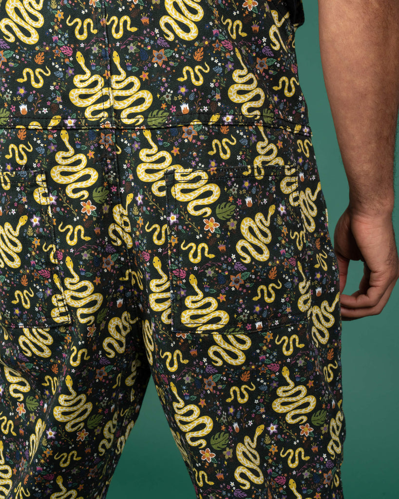 Close-up of the back of Run & Fly dungarees featuring a serpent garden print. The pattern shows yellow snakes intertwined with vibrant botanical elements like flowers and leaves, set against a black background. Ethical slow fashion, unisex style, and eco-friendly fabric choices make these a standout. Model showcases rear view with intricate snake detailing.