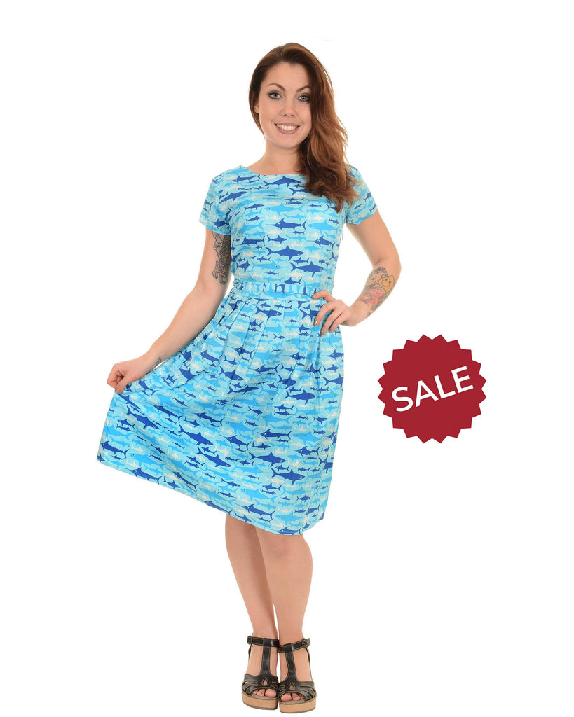 Model is stood in front of a white studio background wearing the jaws tea party dress with black strap sandal heels. They are facing the camera posing with one hand on their hip and the other holding out the bottom of the dress and smiling.